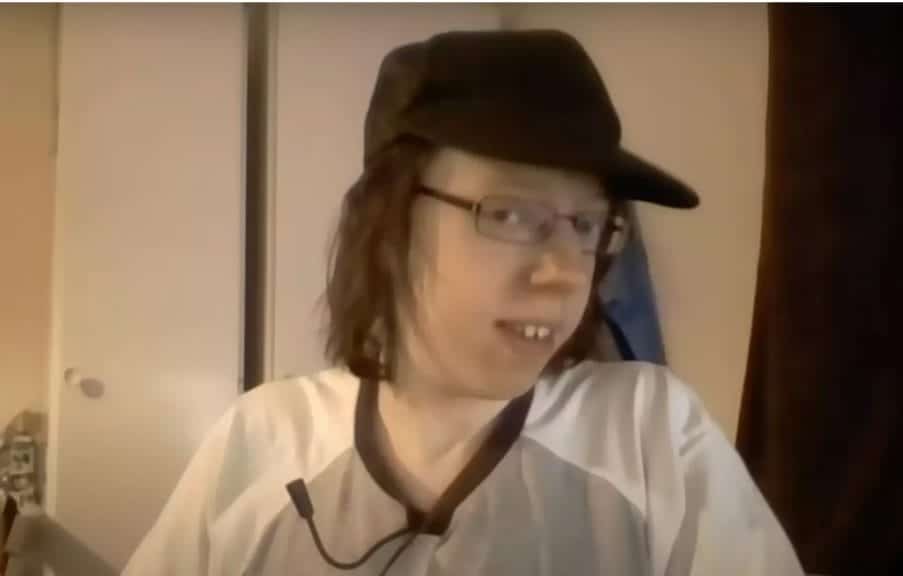 Video of man recording him self. He has a black hat and glasses with long straight hair and a white and grey shirt.